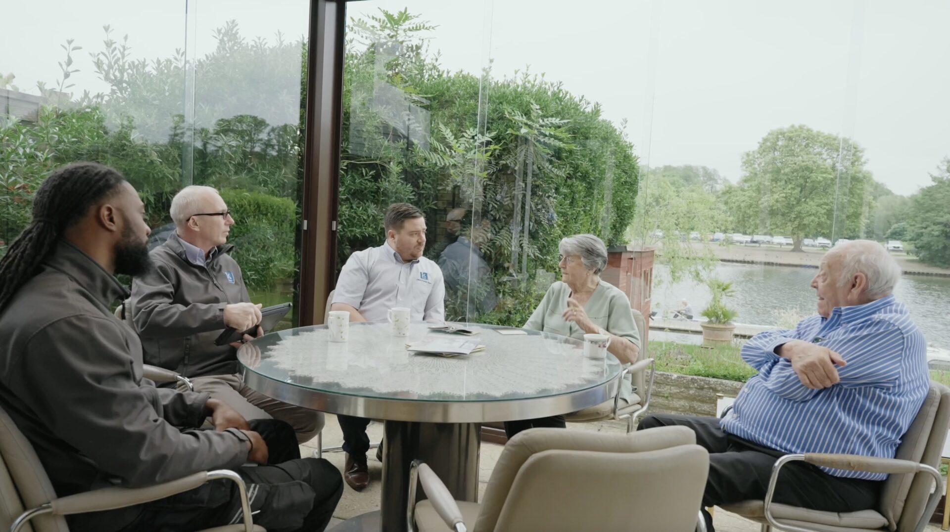 Header image for blog titled: 'A Customer Journey with Kudos Stairlifts - Mr & Mrs Smith'. Screenshot taken from customer video, showing customers sat around table at home with member of Kudos Stairlifts' Client Focus Team and stairlift engineers.